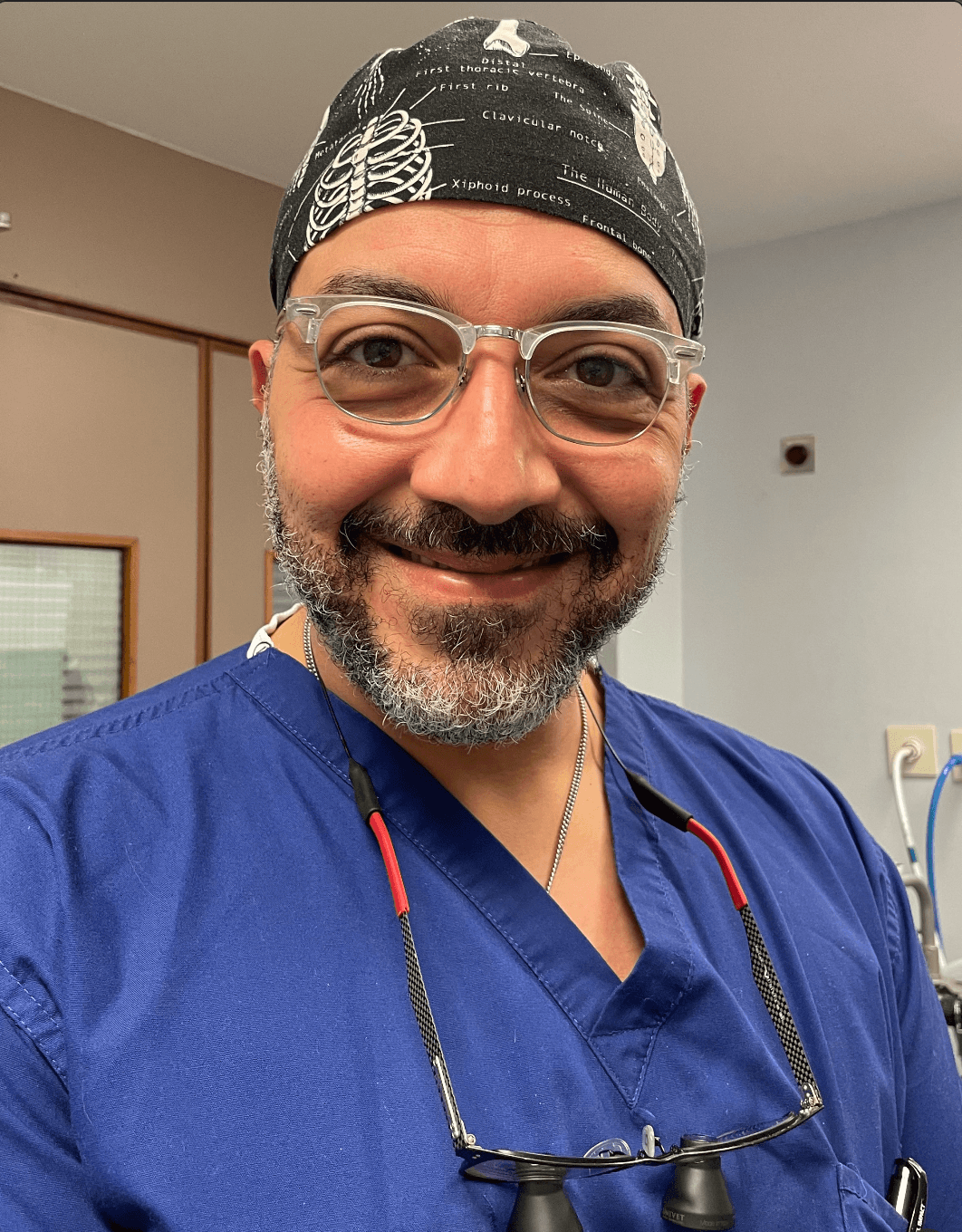 Mr Sherif Elshazly, smiling, in surgical scrubs and an anatomy-print cap, with loupes around his neck